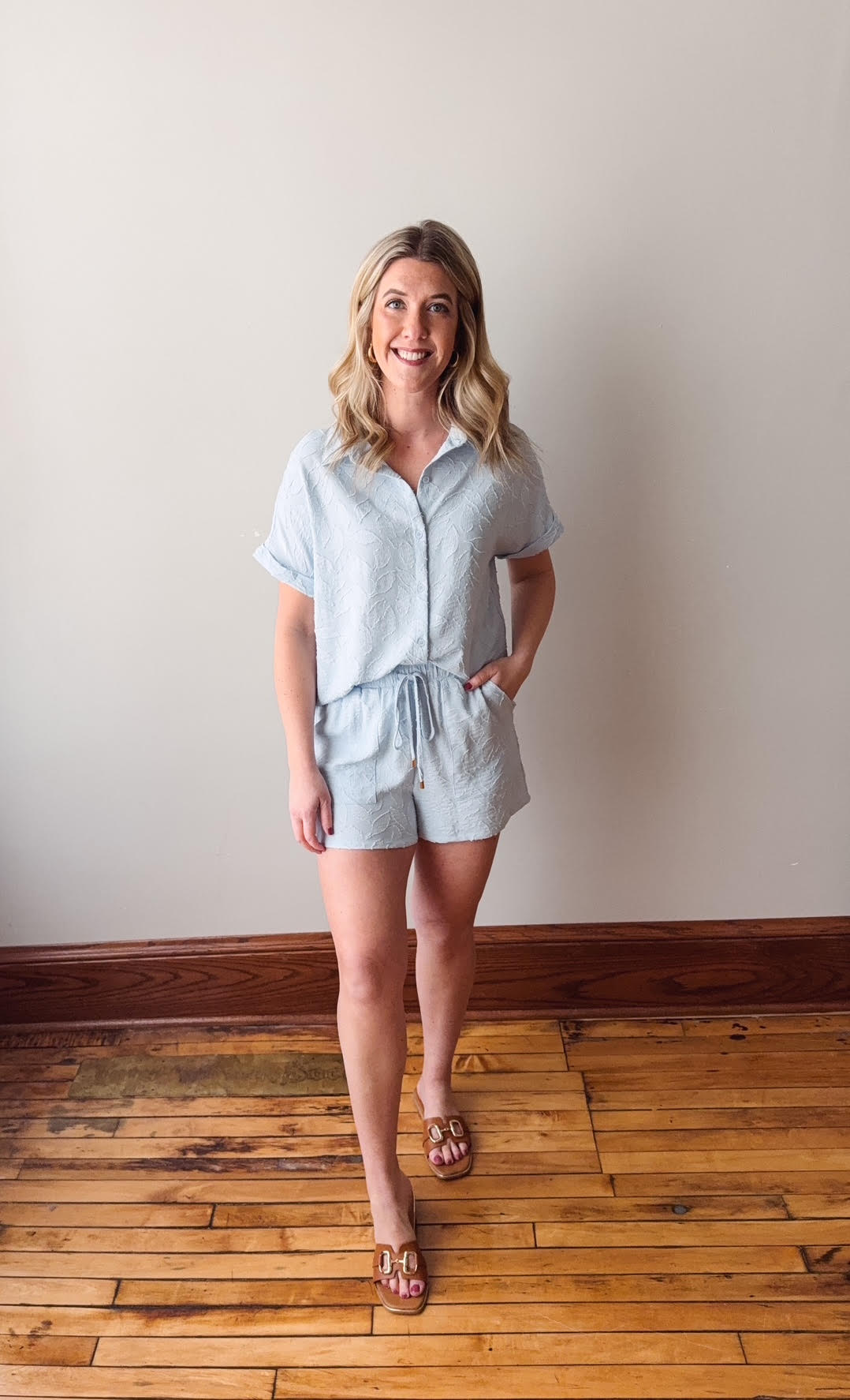 Woman wearing a light blue romper standing on a wooden floor against a plain wall.