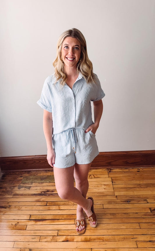 Woman wearing a light blue romper standing on a wooden floor with a white wall background