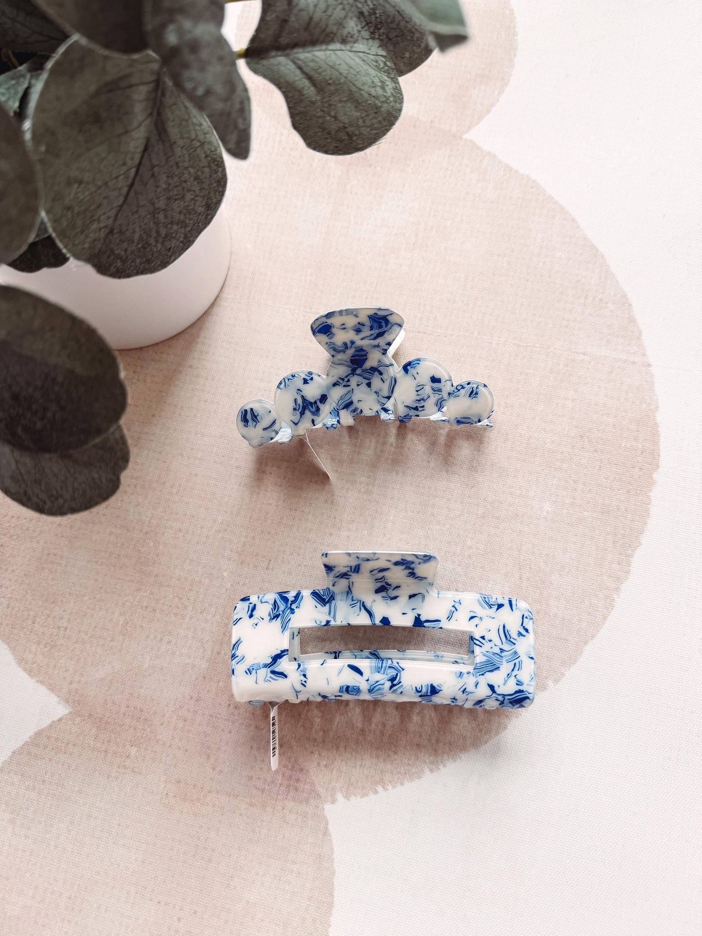 Two blue floral hair clips on a beige surface with a plant in the background