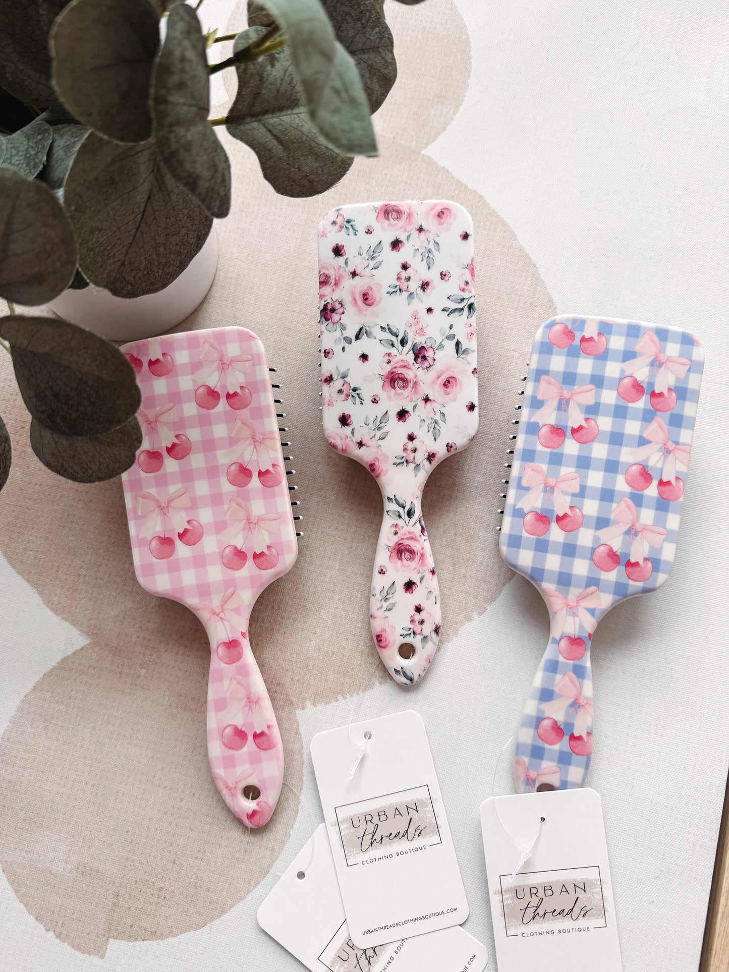 Three floral-patterned hairbrushes on a light surface with a plant in the background.