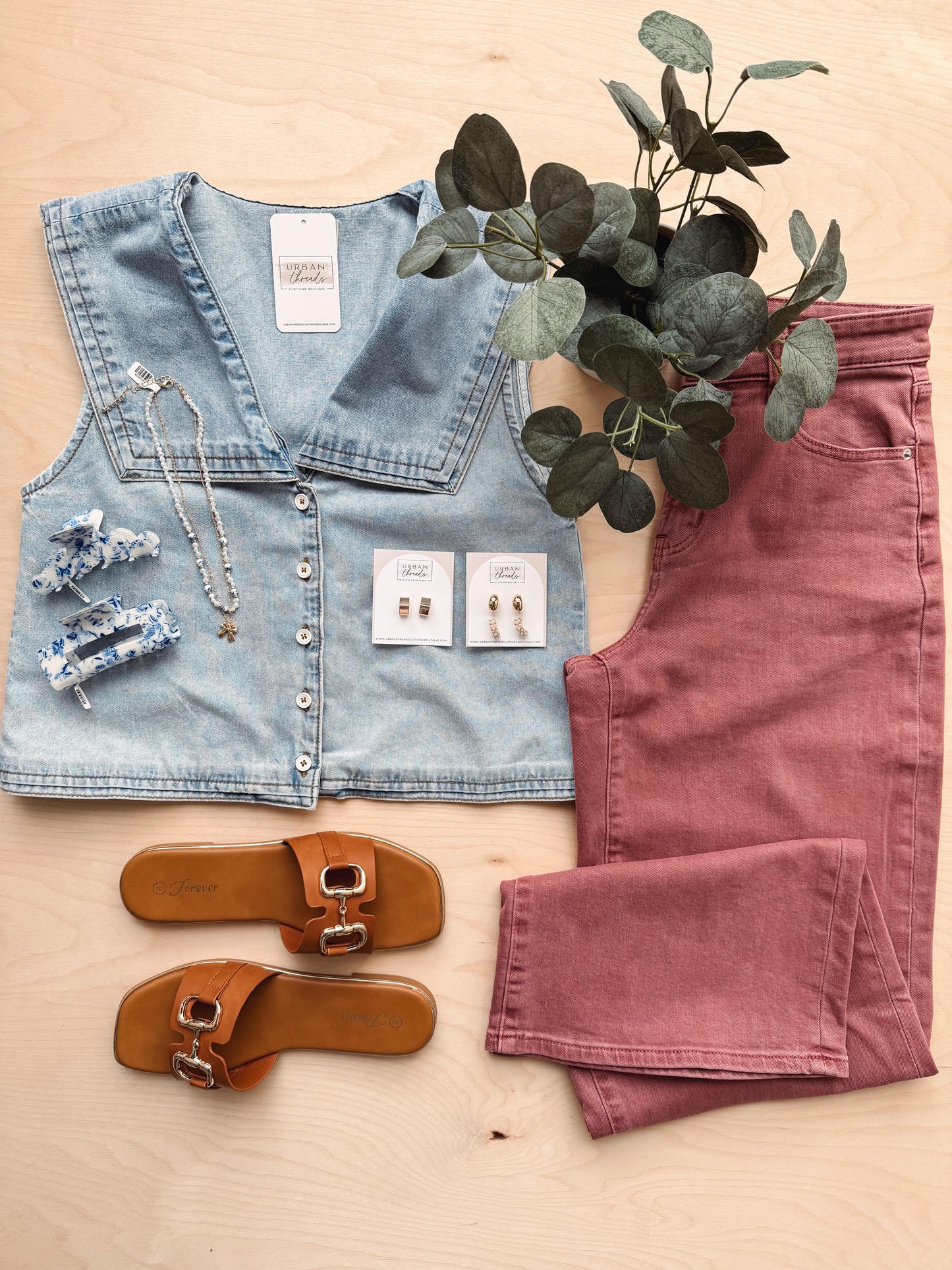 Denim vest, pink pants, brown sandals, and jewelry on a wooden surface with a plant.