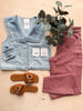 Denim vest, pink pants, brown sandals, and jewelry on a wooden surface with a plant.