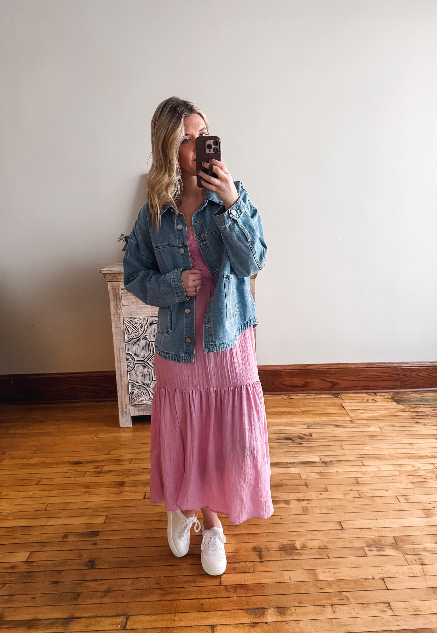 Woman taking a mirror selfie wearing a denim jacket and pink dress on a wooden floor.