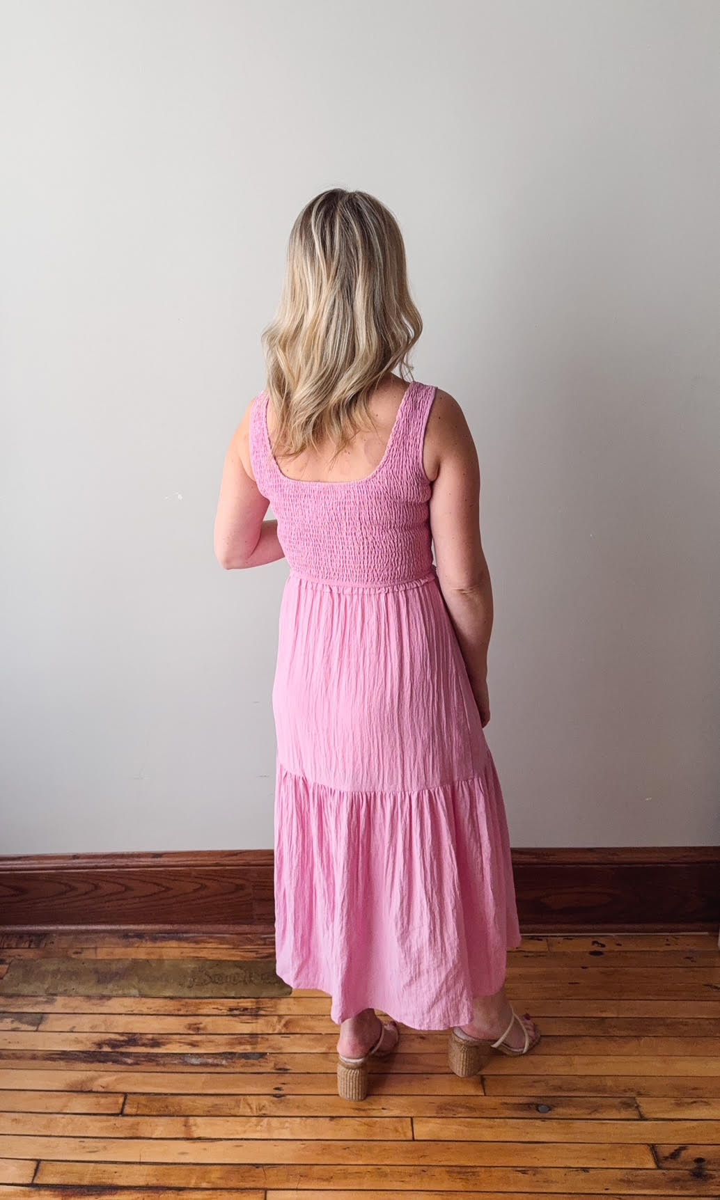 Woman wearing a pink dress standing in a room with wooden flooring and a white wall.