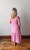 Woman wearing a pink dress standing in a room with wooden flooring and a white wall.