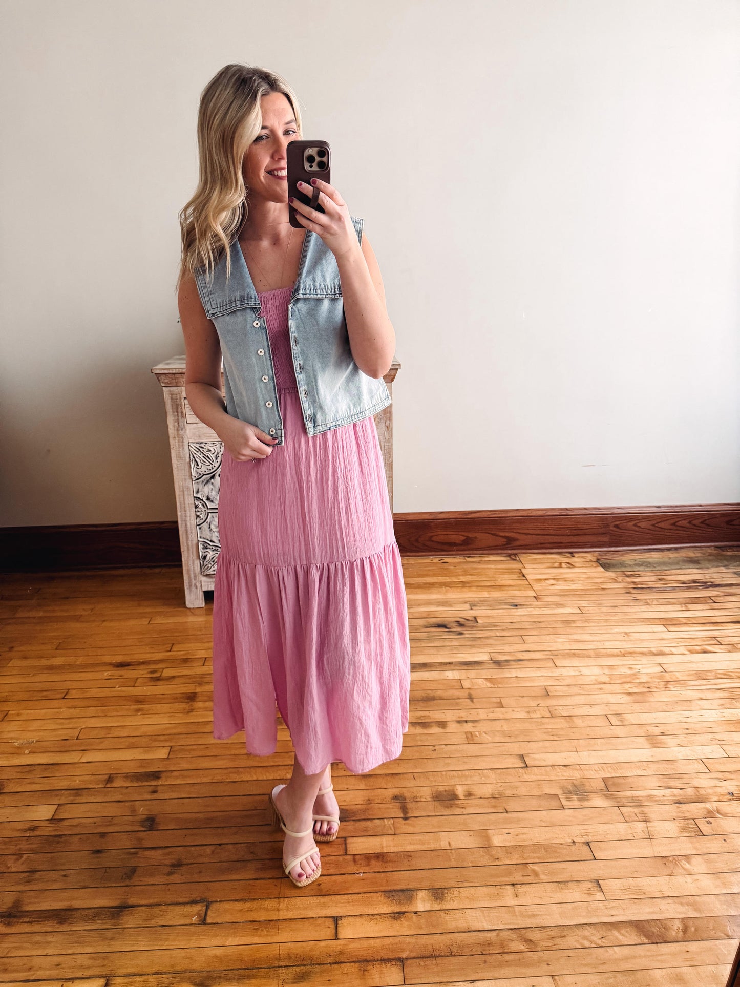 Woman taking a mirror selfie wearing a pink dress and denim vest on a wooden floor.
