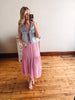 Woman taking a mirror selfie wearing a pink dress and denim vest on a wooden floor.