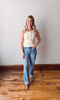 Woman wearing a sleeveless top and jeans standing on a wooden floor with a white wall background