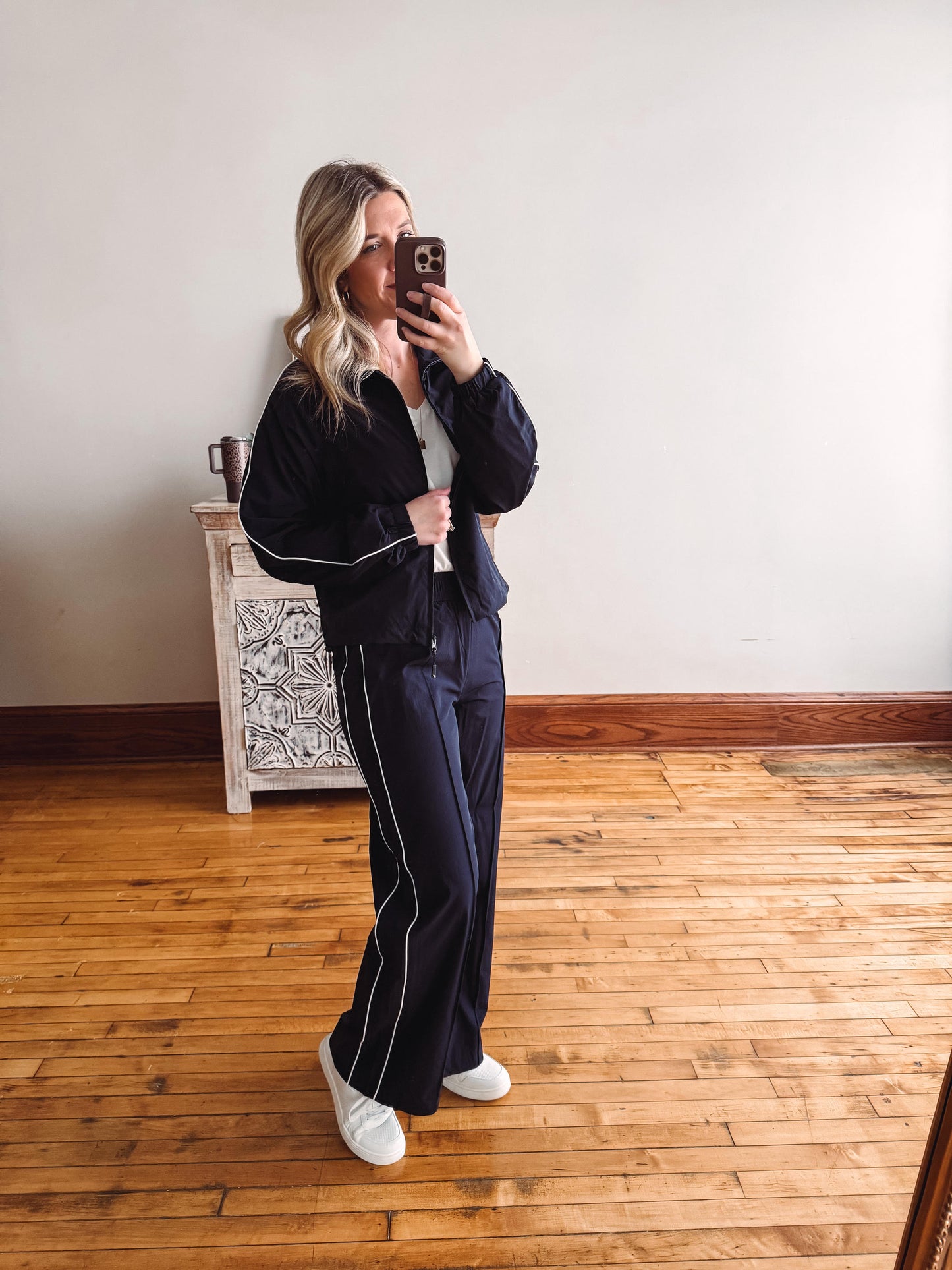 Person taking a mirror selfie wearing a black tracksuit with white stripes on a wooden floor.