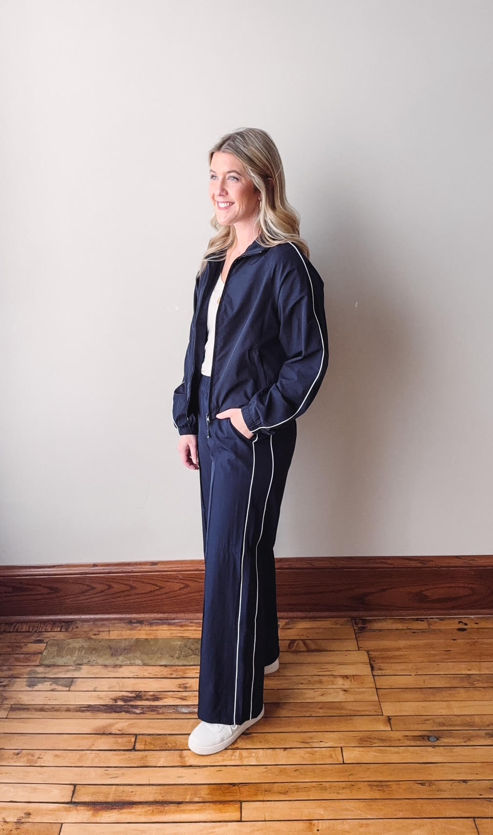 Woman wearing a navy tracksuit with white piping standing on a wooden floor against a plain wall.