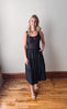 Woman wearing a black sleeveless dress standing on a wooden floor against a plain wall.