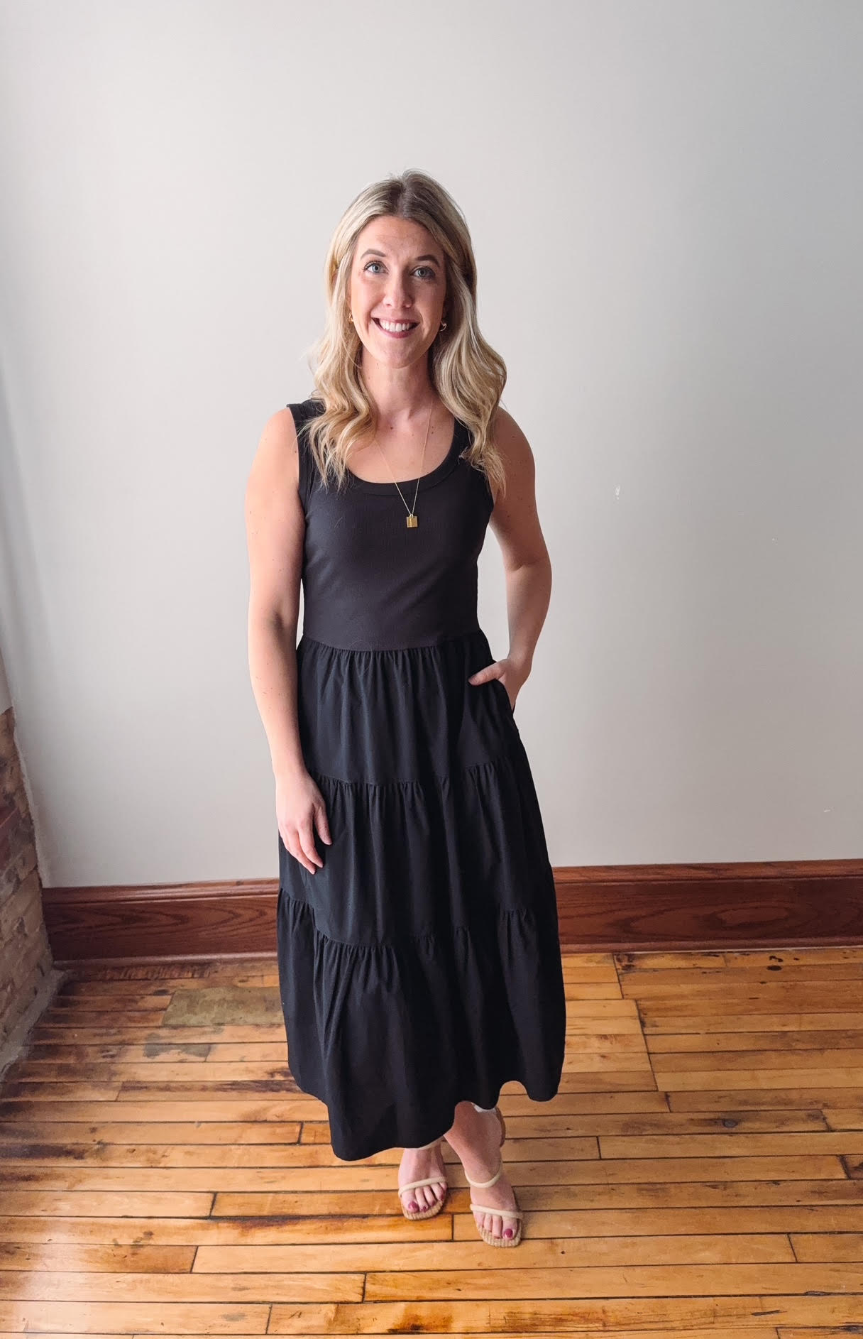 Woman wearing a black sleeveless dress standing on a wooden floor with a white wall background
