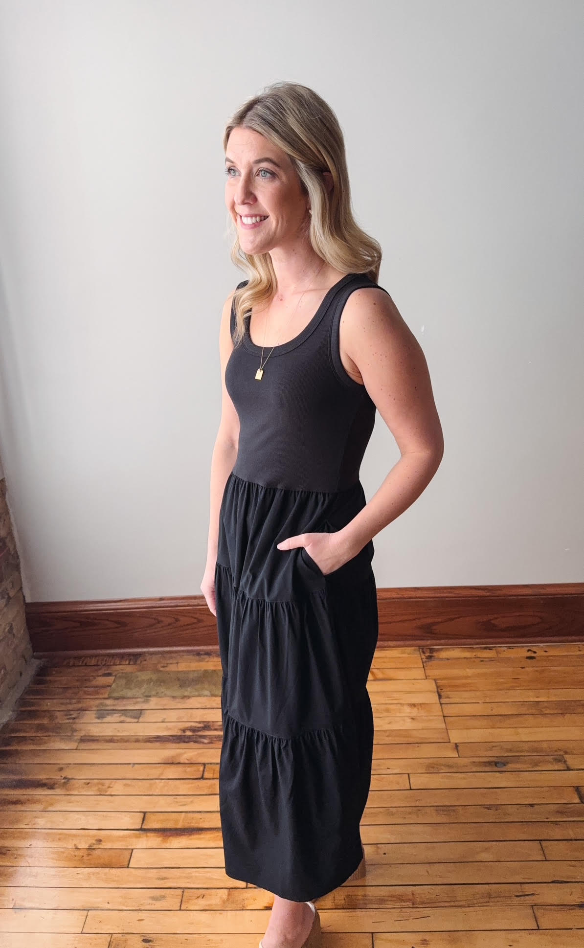 Woman wearing a black dress standing in a room with wooden flooring and a white wall.