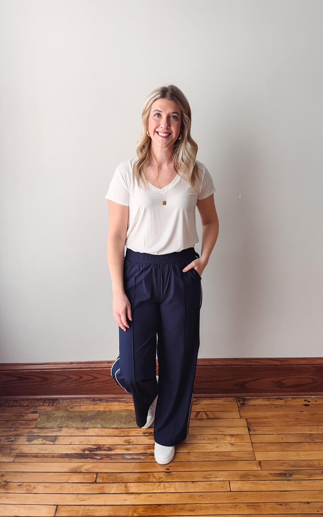 Woman wearing a white t-shirt and navy pants standing on a wooden floor with a plain wall background