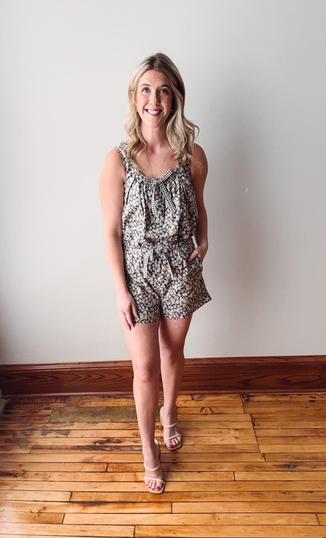 Woman wearing a patterned romper standing on a wooden floor with a white wall background
