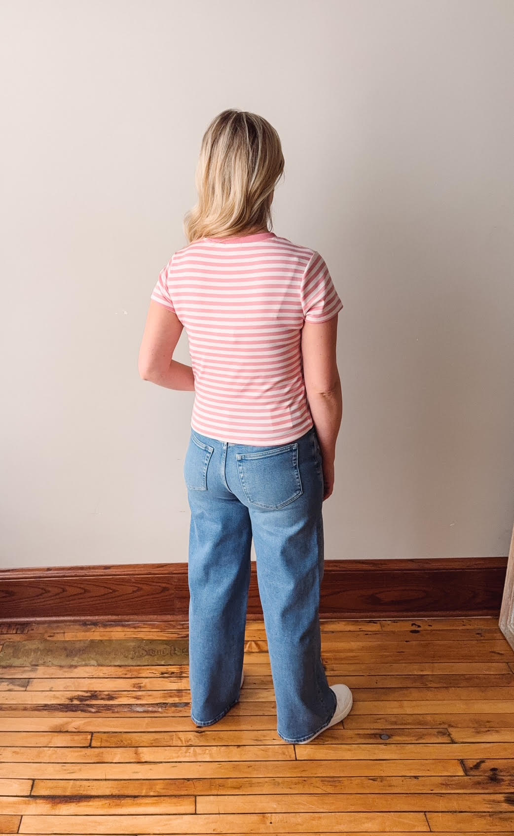 Person wearing a striped shirt and jeans standing on a wooden floor against a plain wall.
