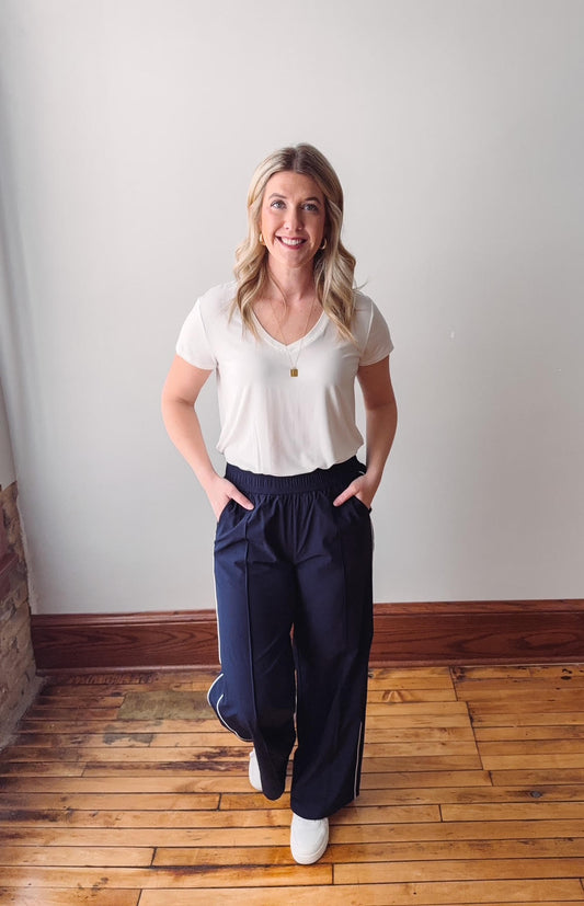 Woman wearing a white t-shirt and navy pants standing on a wooden floor against a white wall.