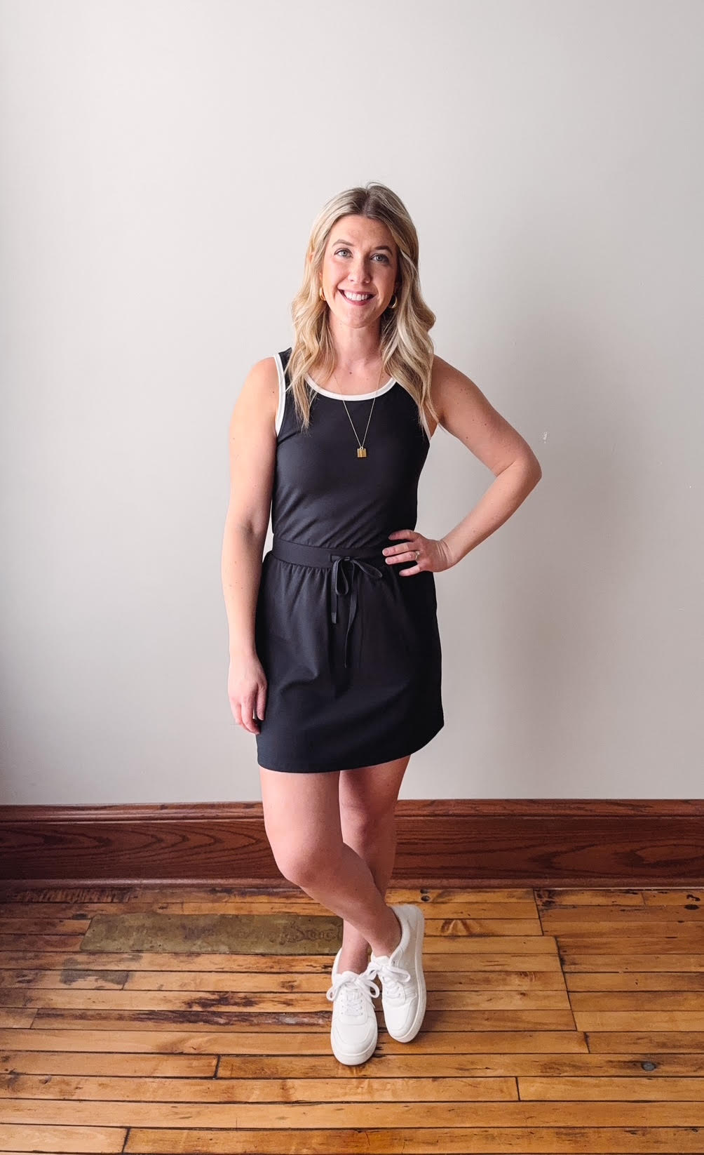 Woman wearing a black dress standing on a wooden floor with a white wall background