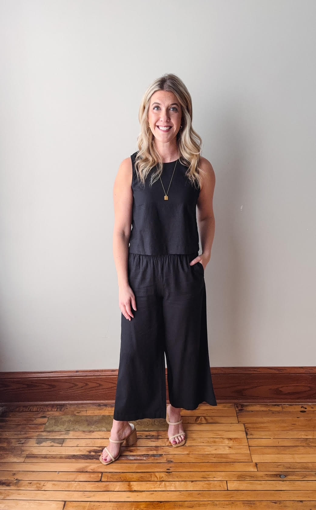 Woman wearing a black sleeveless top and wide-leg pants standing on a wooden floor against a white wall.