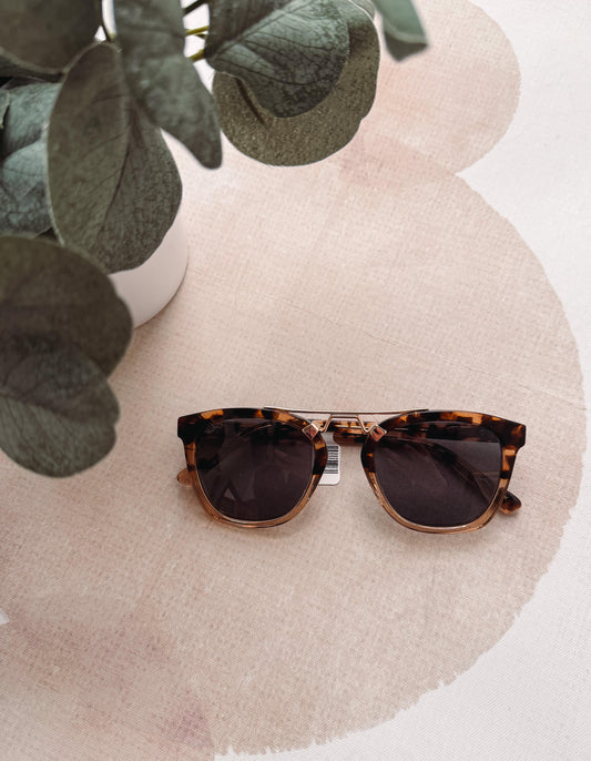 Tortoiseshell sunglasses on a beige surface with green leaves in the corner