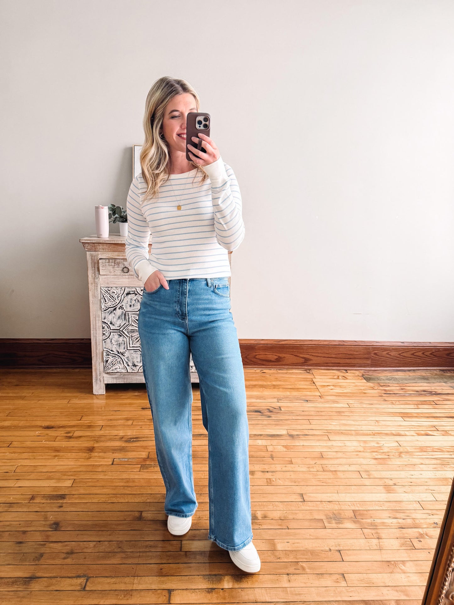 Woman taking a mirror selfie wearing a striped sweater and blue jeans in a room with wooden flooring.