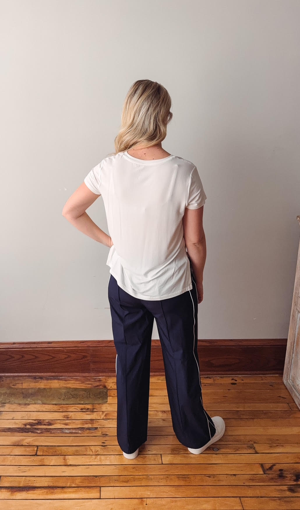 Person wearing a white t-shirt and navy pants standing in a room with wooden flooring and a plain wall.