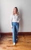 Woman wearing a striped sweater and blue jeans standing on a wooden floor against a white wall.