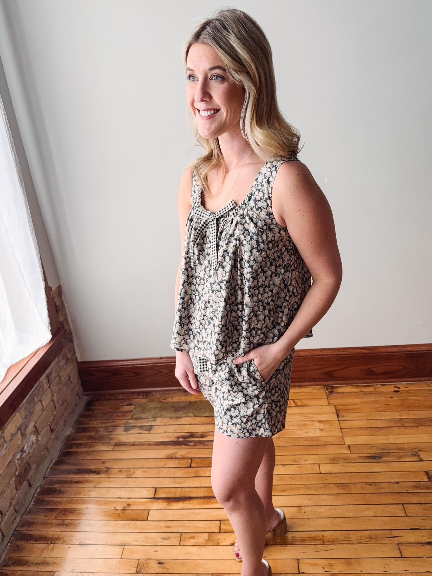 Woman in a patterned dress standing in a room with wooden flooring and a white wall.