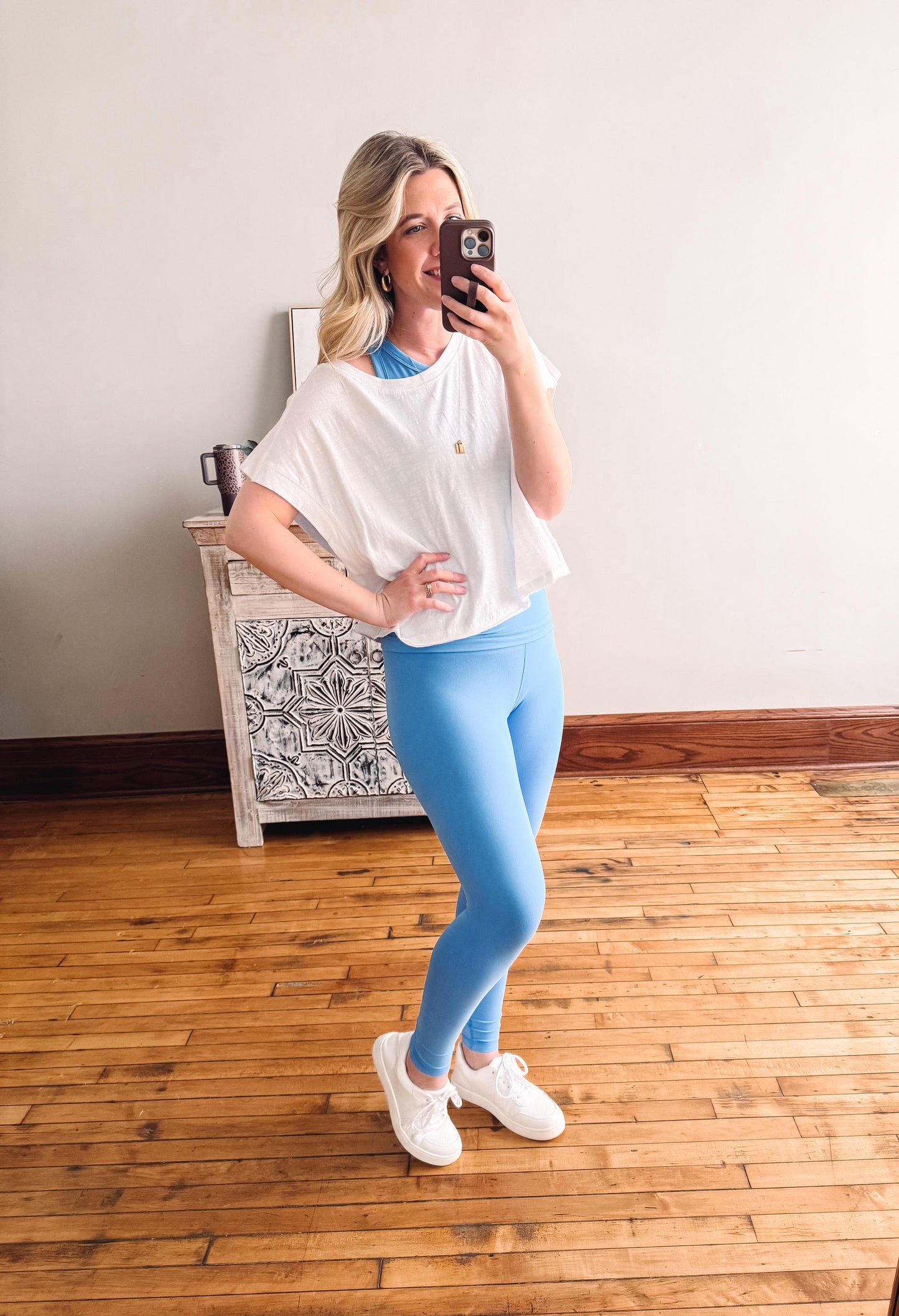 Woman taking a mirror selfie wearing a white top and blue leggings in a room with wooden flooring.