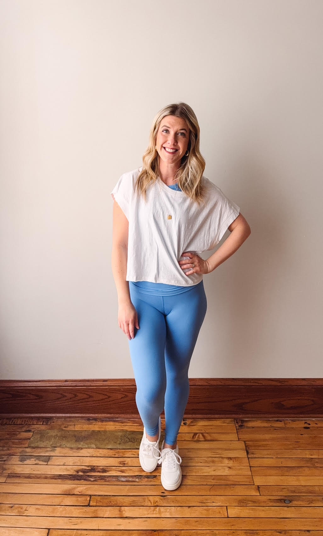 Woman wearing a white shirt and blue leggings standing on a wooden floor against a plain wall.