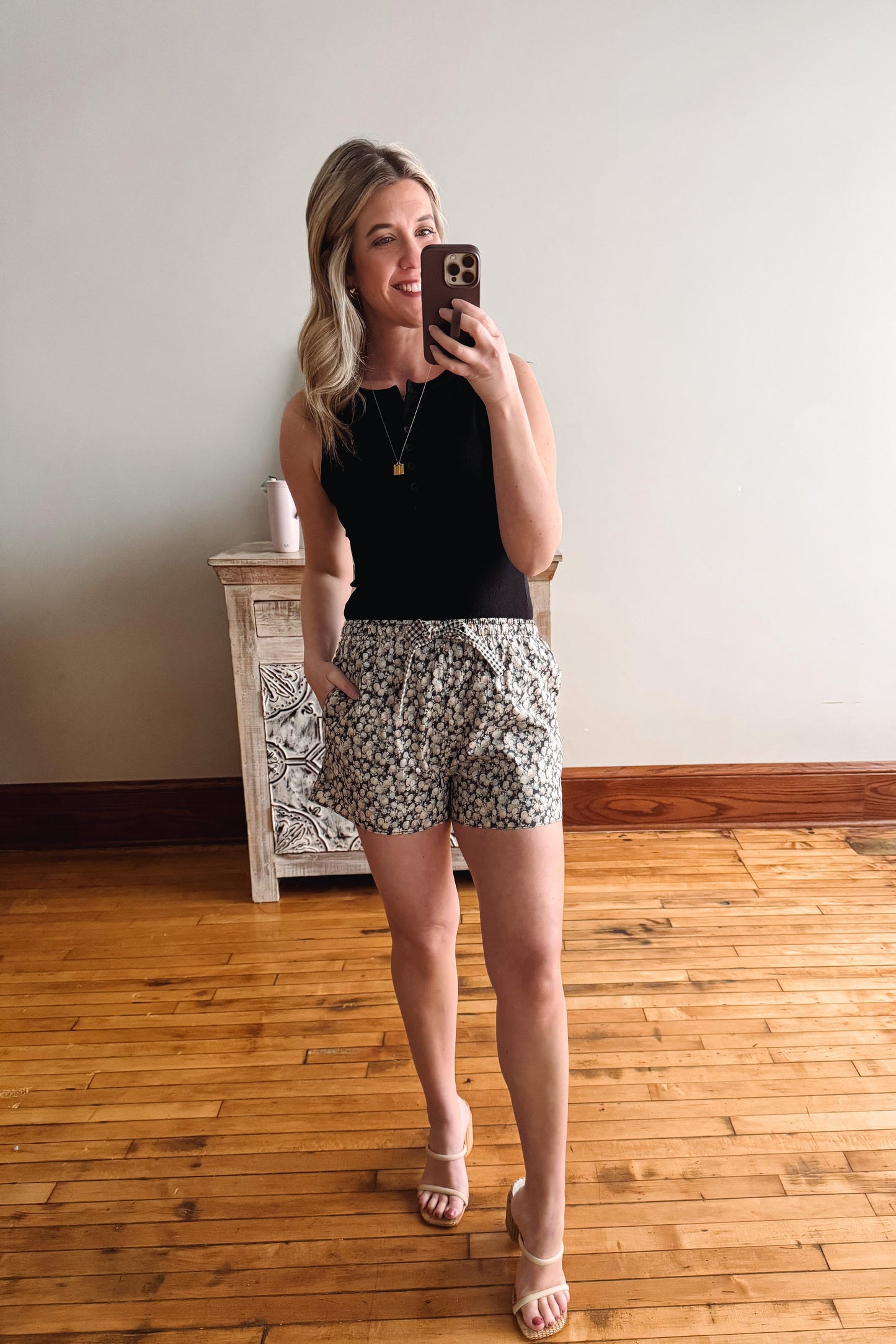 Woman taking a mirror selfie wearing a black top and floral shorts in a room with wooden flooring.