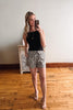 Woman taking a mirror selfie wearing a black top and floral shorts in a room with wooden flooring.