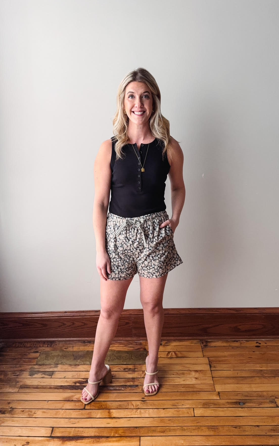Woman wearing a black top and patterned shorts standing on a wooden floor with a white wall background