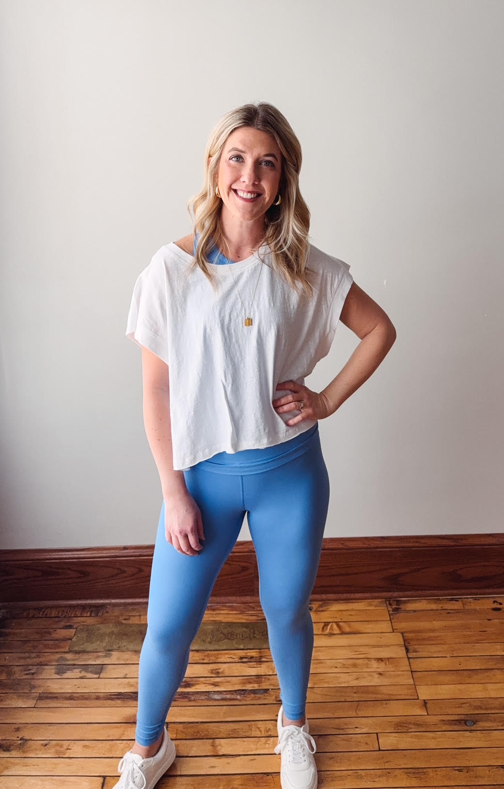 Woman wearing a white top and blue leggings standing on a wooden floor.