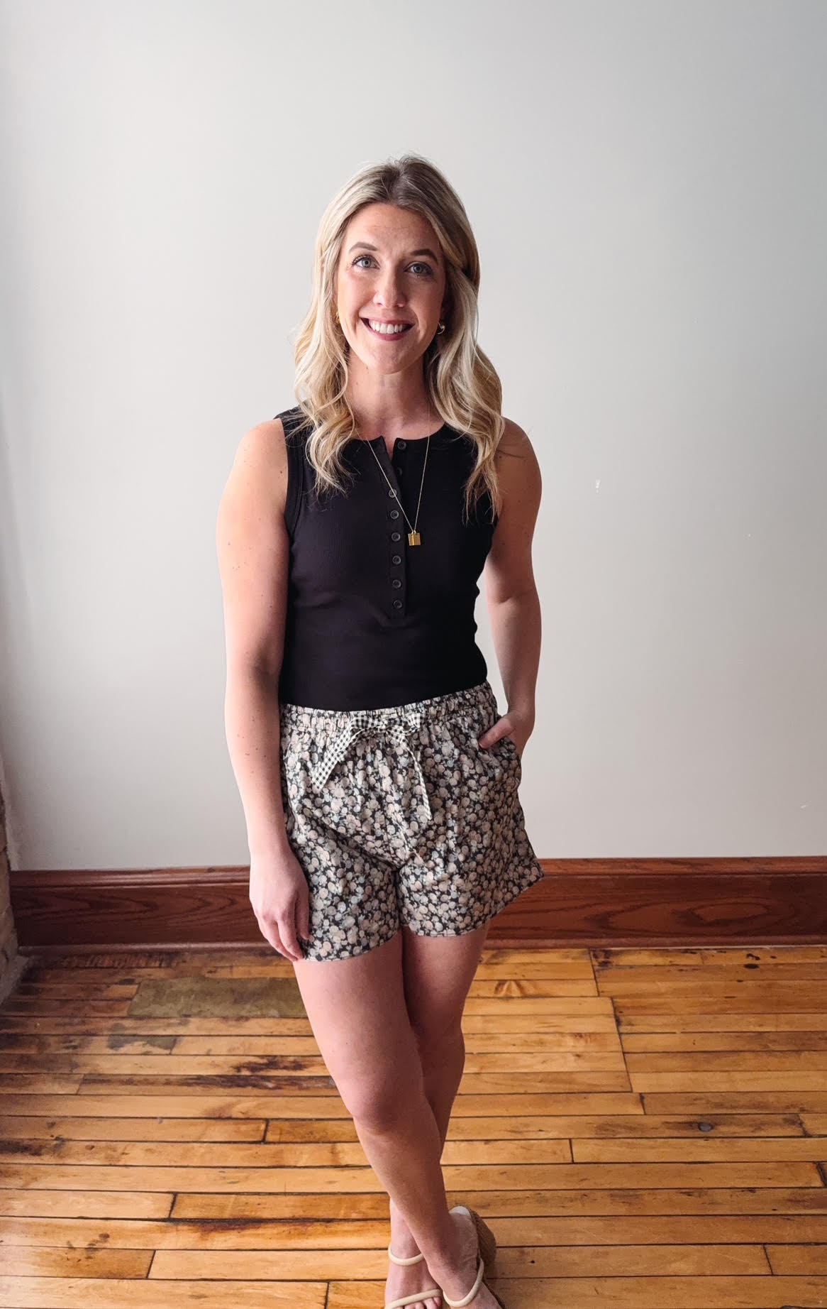 Woman wearing a black sleeveless top and patterned shorts standing on a wooden floor.