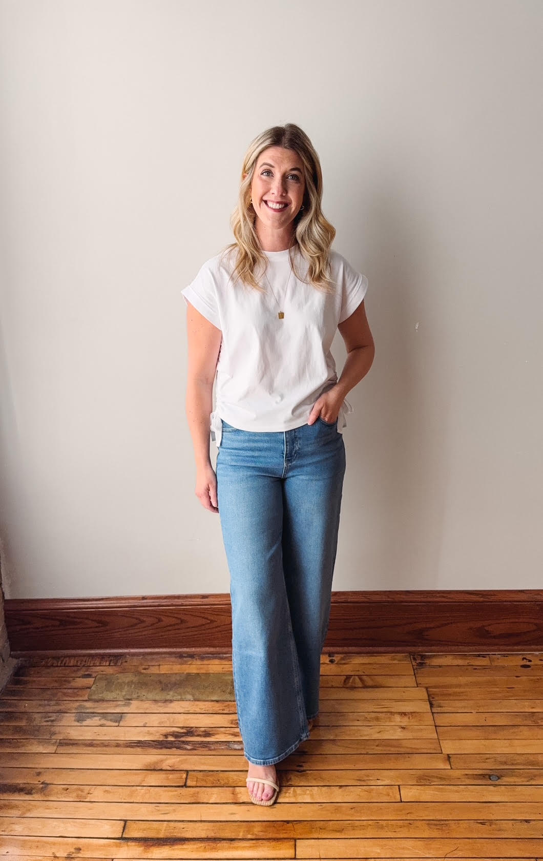 Woman wearing a white t-shirt and blue jeans standing against a plain wall.