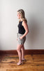 Woman wearing a black top and patterned shorts standing on a wooden floor against a white wall.