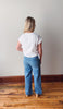 Person wearing a white t-shirt and blue jeans standing on a wooden floor with a plain wall background