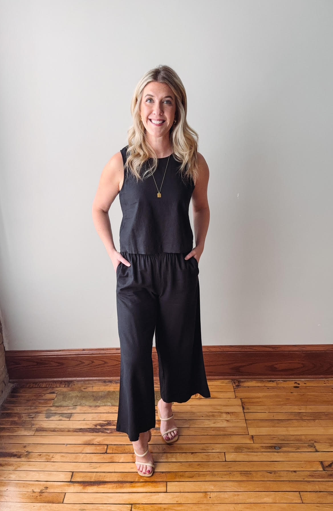 Woman wearing a black sleeveless top and wide-leg pants standing on a wooden floor.