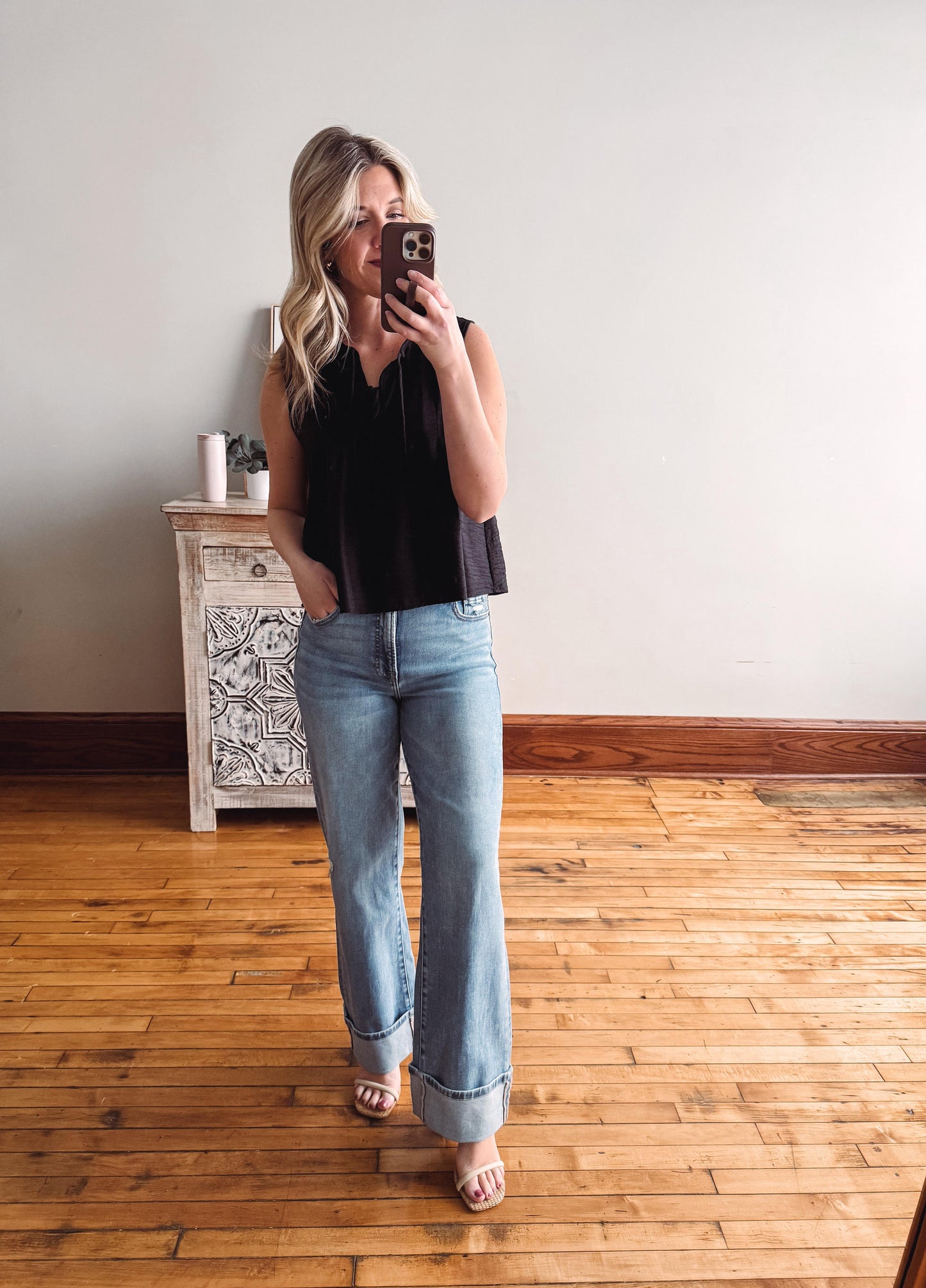 Woman taking a mirror selfie wearing a black sleeveless top and light blue jeans in a room with wooden flooring.