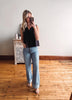 Woman taking a mirror selfie wearing a black sleeveless top and light blue jeans in a room with wooden flooring.
