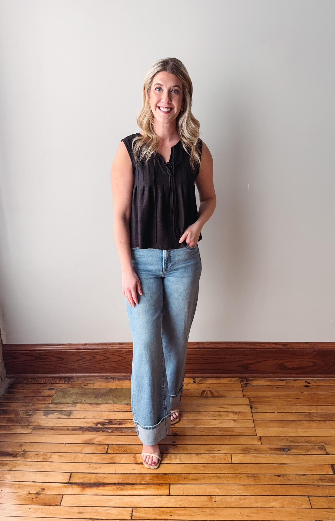 Woman wearing a black sleeveless top and blue jeans standing on a wooden floor.