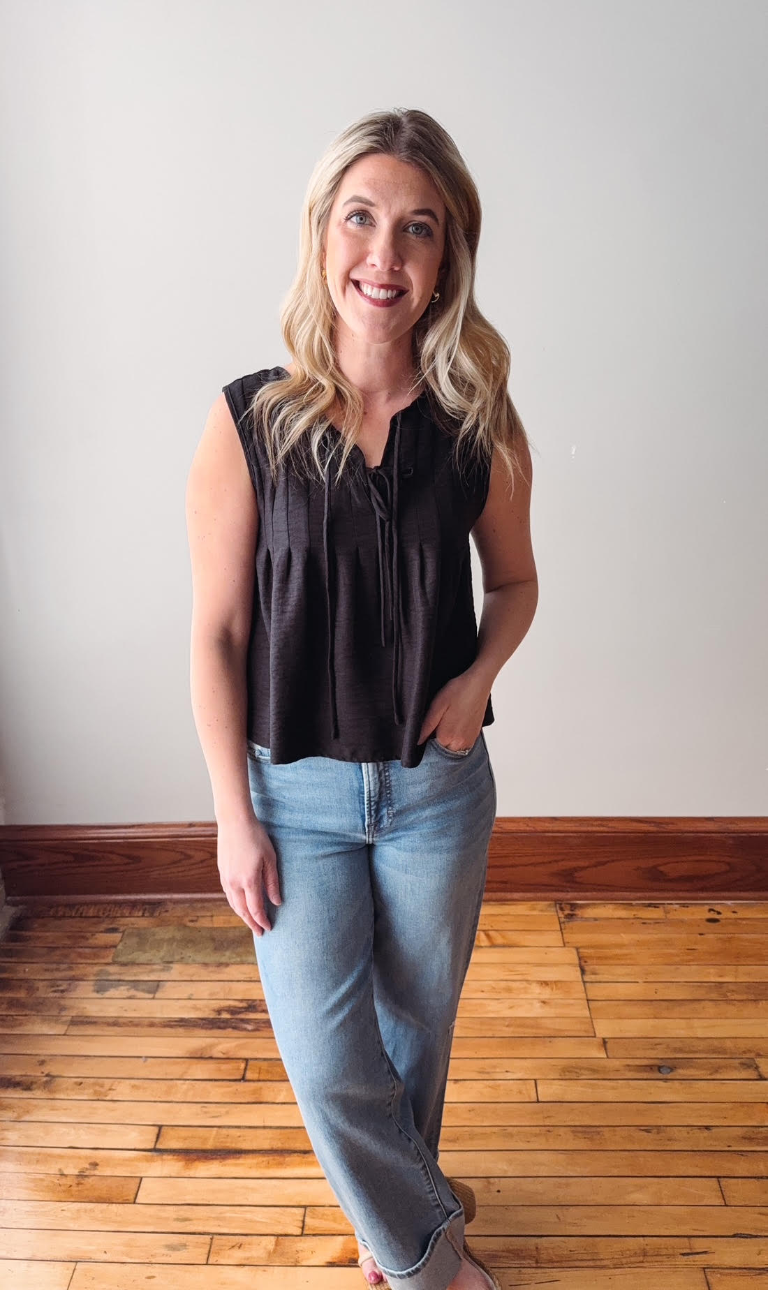Woman wearing a black sleeveless top and blue jeans standing on a wooden floor.