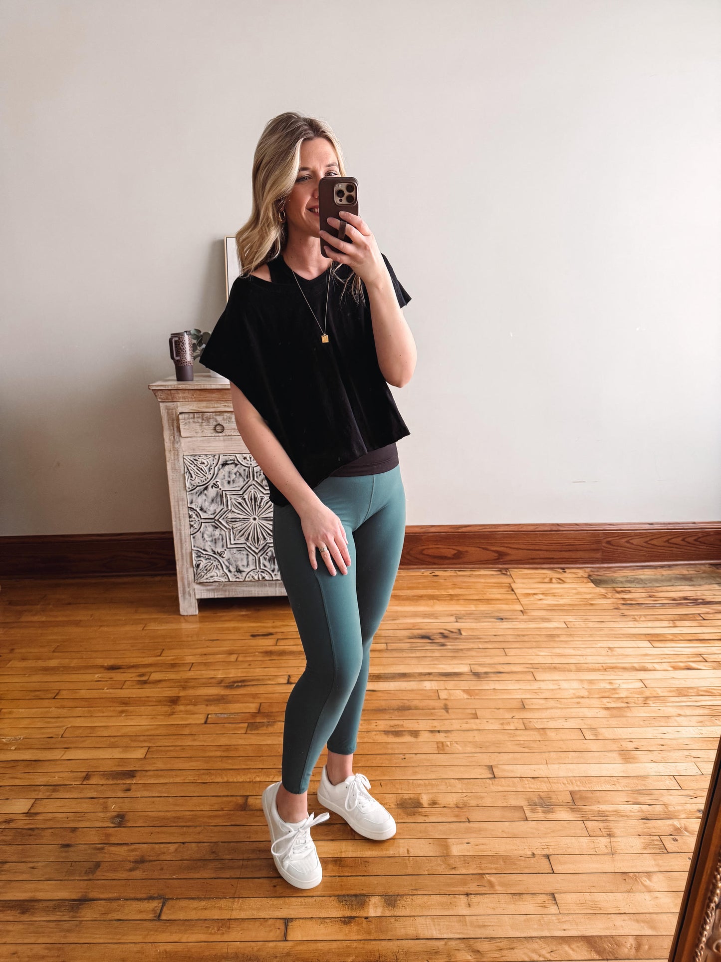 Woman taking a mirror selfie wearing a black top and teal leggings in a room with wooden flooring.