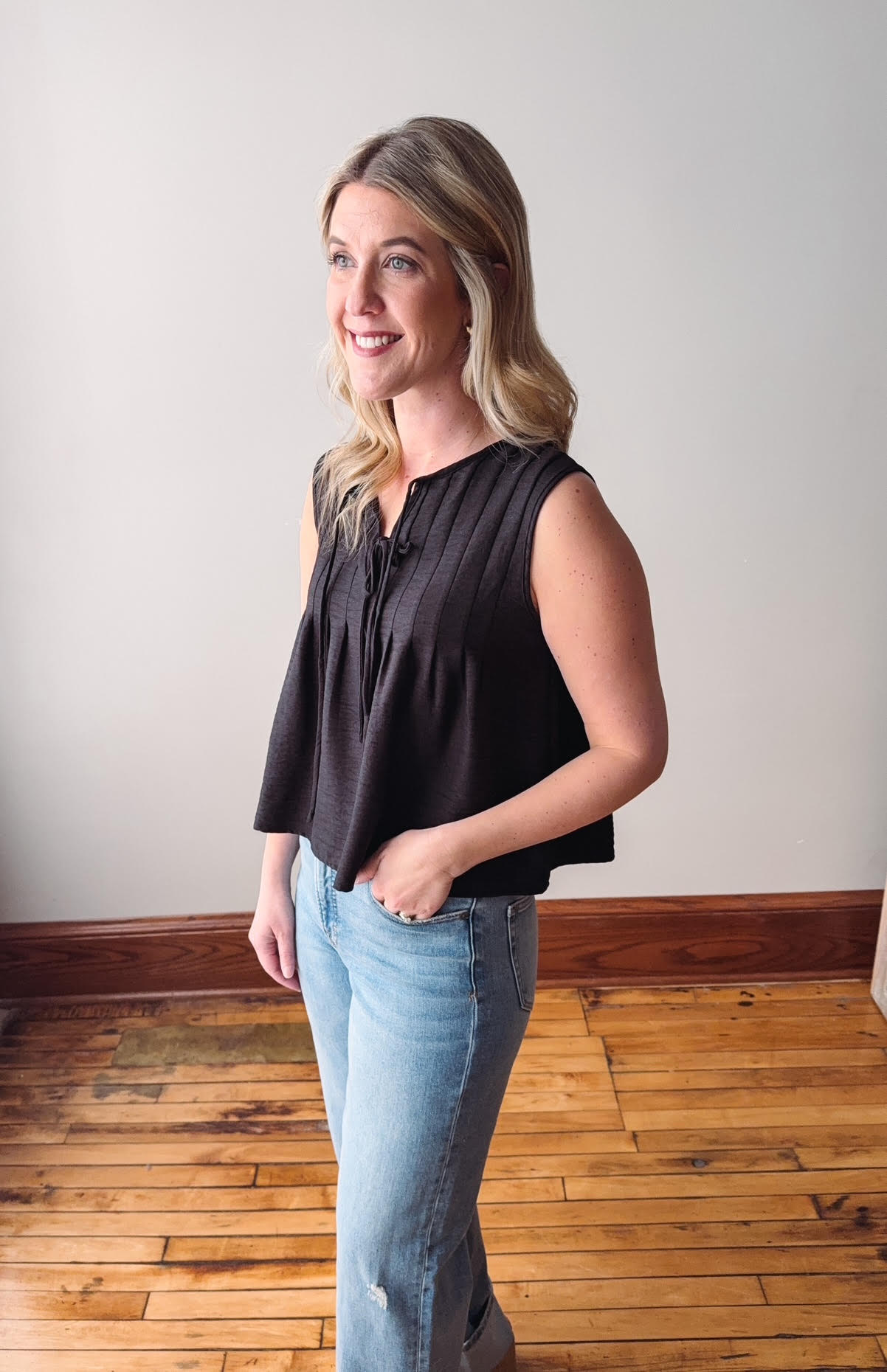 Woman wearing a black sleeveless top and light blue jeans standing in a room with wooden flooring.