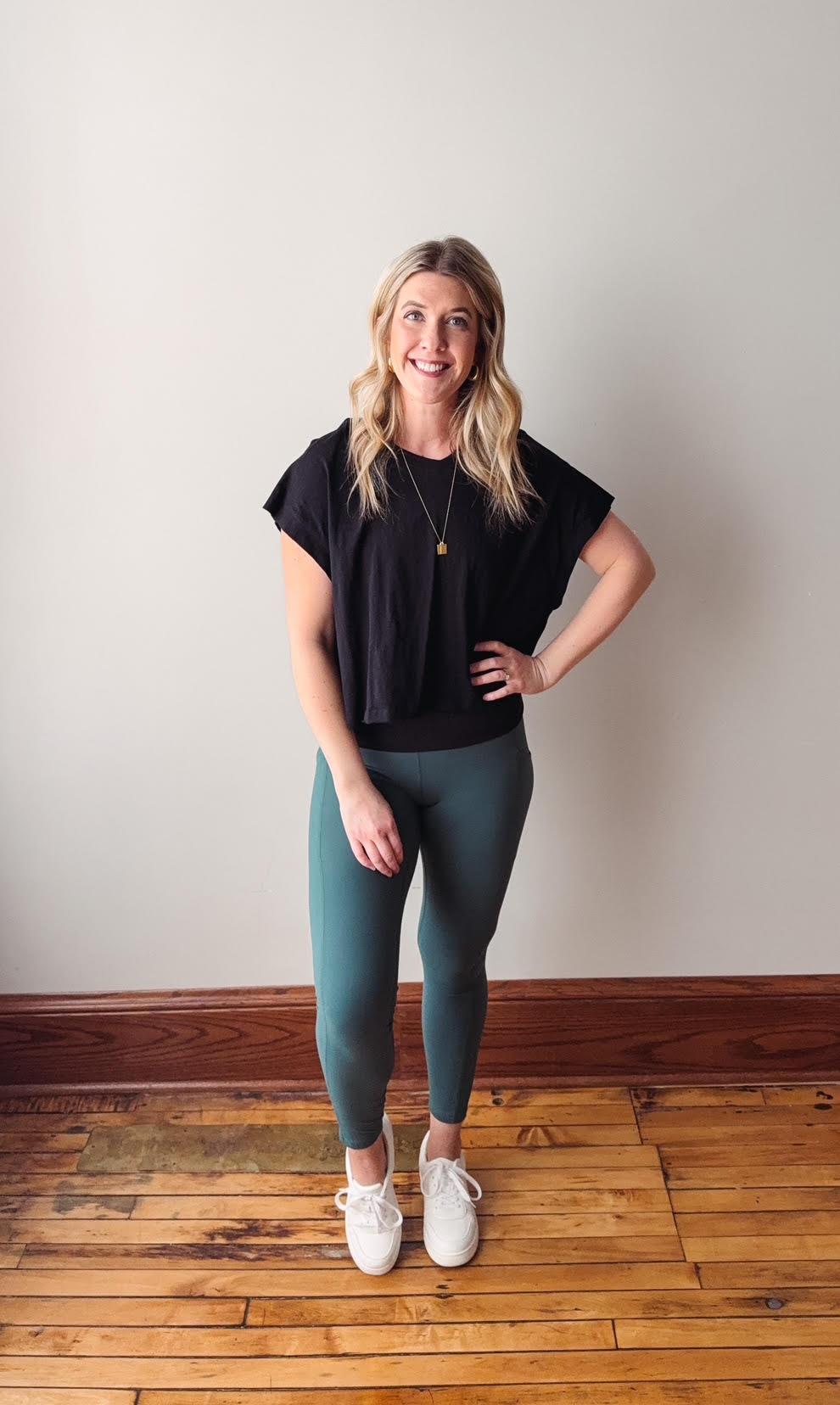 Woman wearing a black top and green leggings standing on a wooden floor with a white wall background