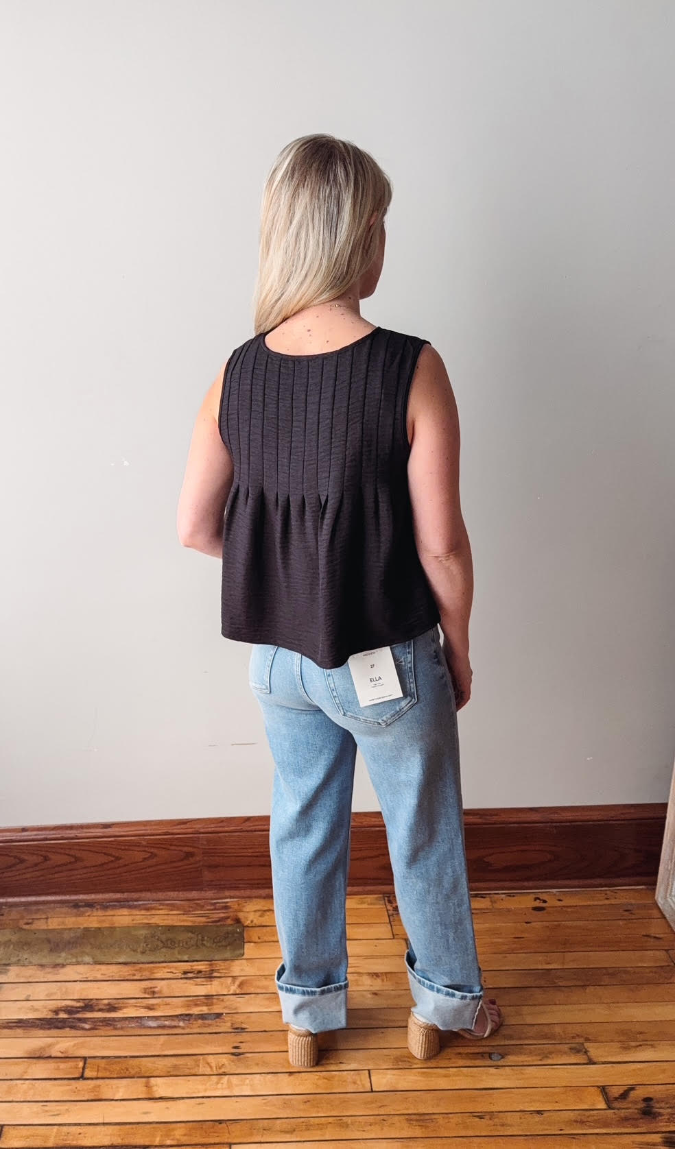 Person wearing a black sleeveless top and light blue jeans standing on a wooden floor with a white wall background.