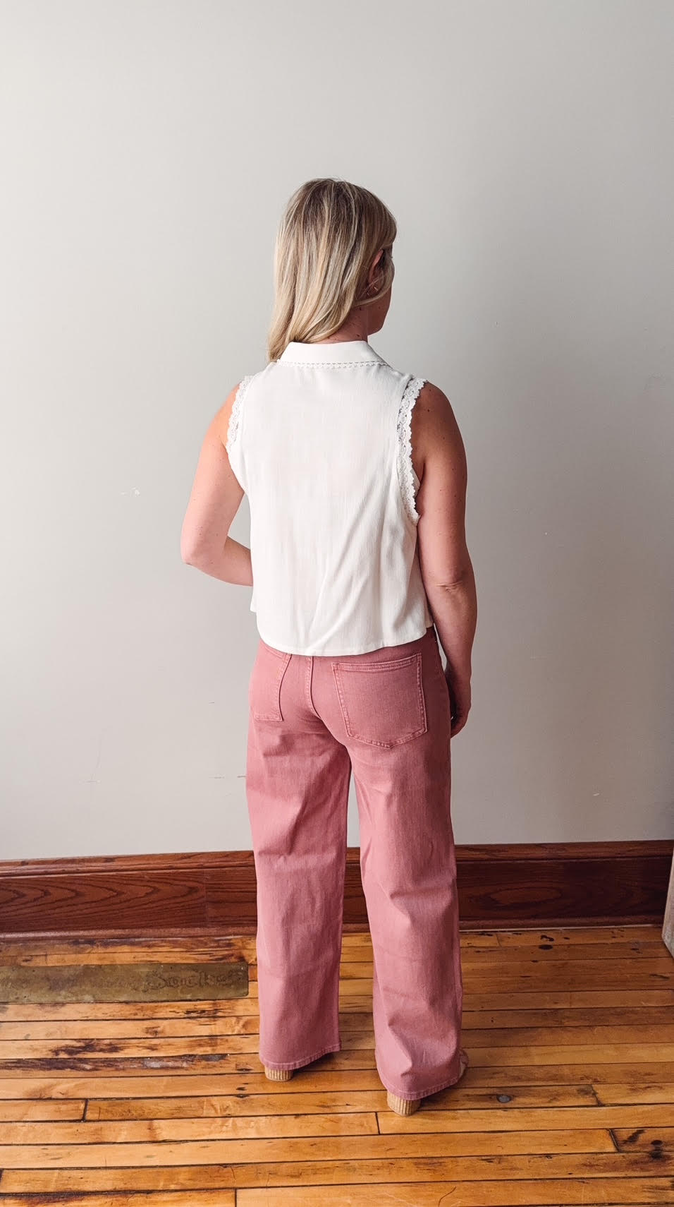 Person wearing a white sleeveless top and pink pants standing on a wooden floor against a plain wall.