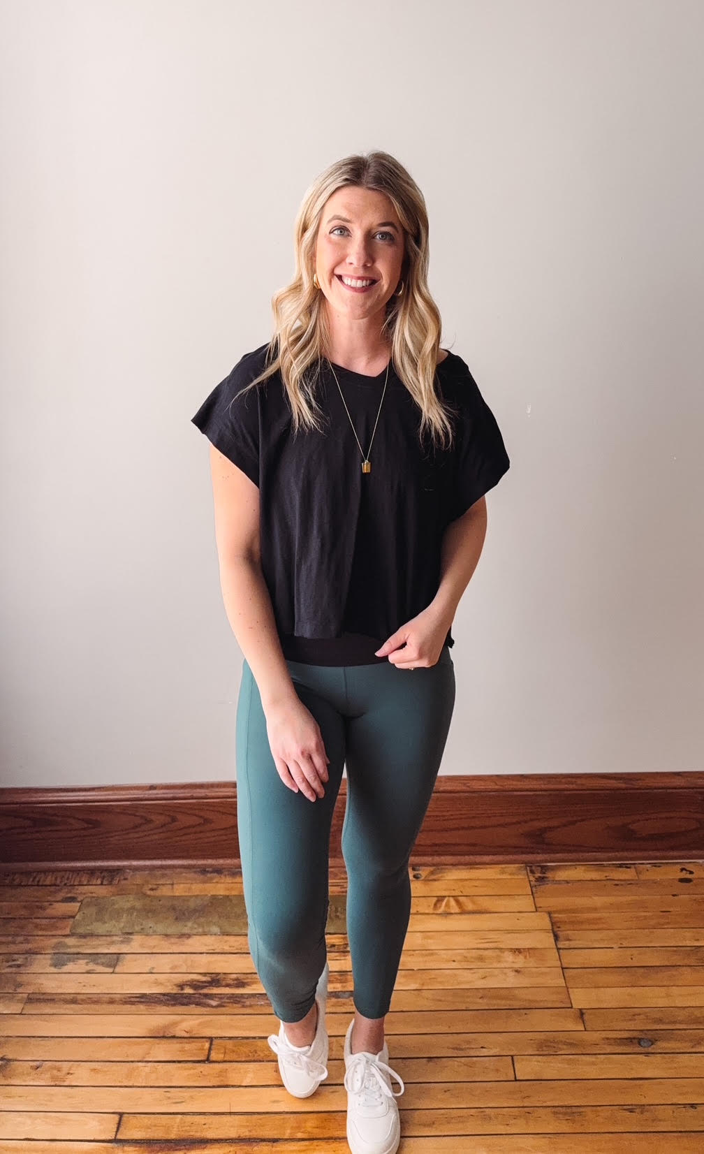 Woman wearing a black top and teal leggings standing on a wooden floor with a plain wall background