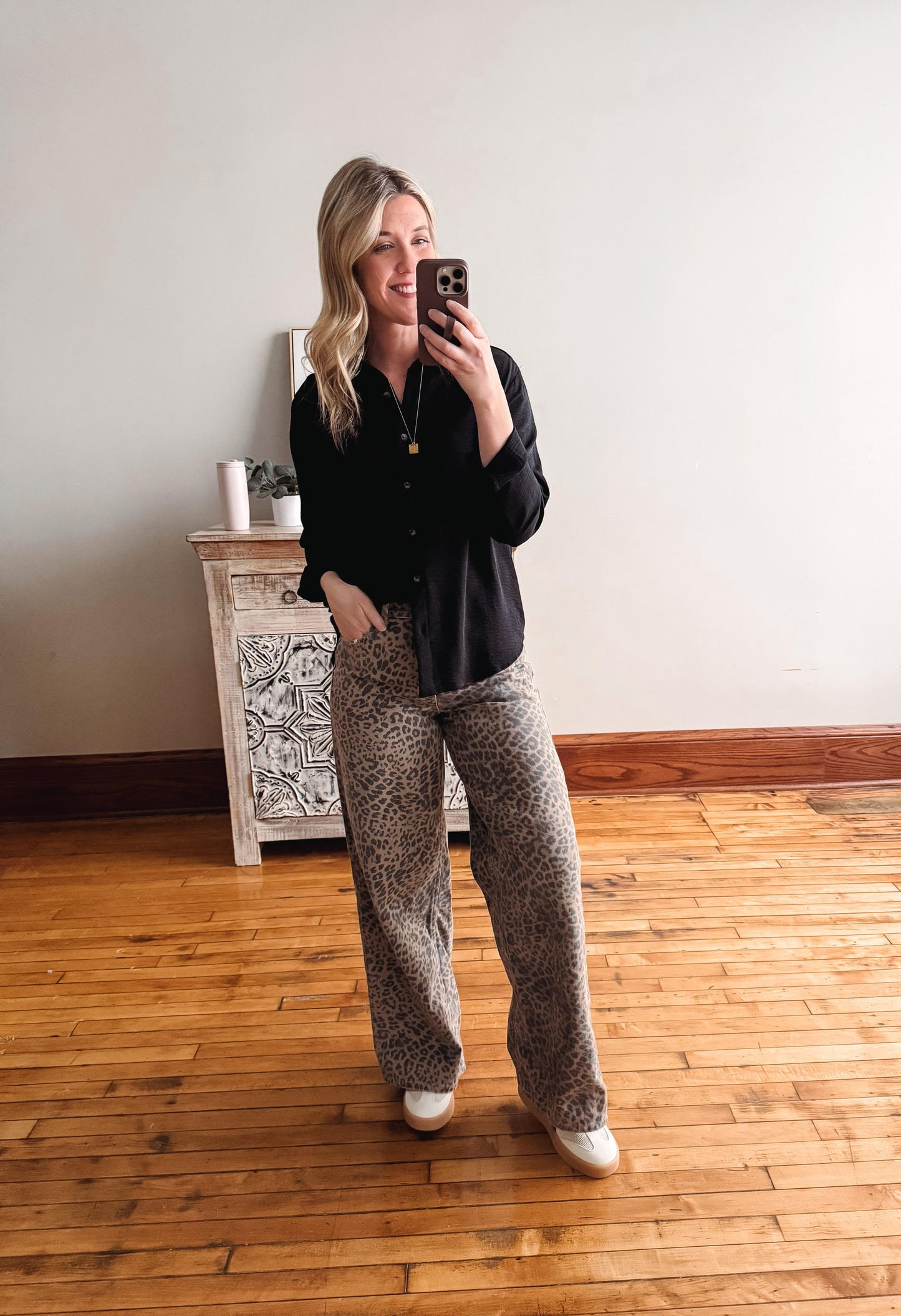Woman taking a mirror selfie wearing a black shirt and patterned pants in a room with wooden flooring.